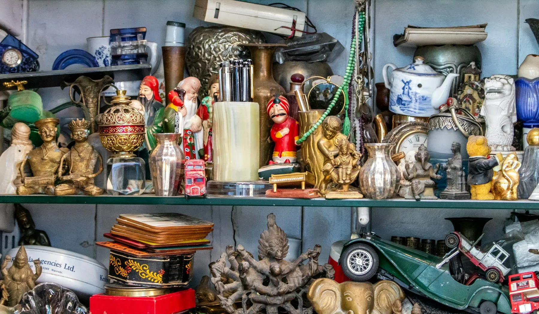 A detailed photograph of a crowded antique shop shelf filled with figurines, brass statues, teapots, clocks, vases, toys